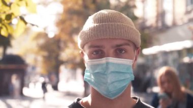 Tıbbi maske takan beyaz, yakışıklı genç adamın yüzü kameraya bakıyor. Sokaktaki adamın portresi. Coronavirus konsepti.
