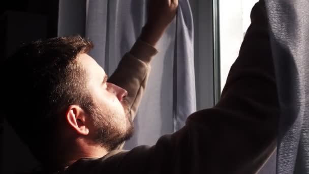 Молодой кавказский мужчина, открывая занавески, глядя в окно наслаждаясь новым днем, чувствует себя отдохнувшим — стоковое видео