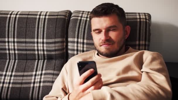 Homem caucasiano lindo sentado no sofá usando smartphone no apartamento moderno mensagem de texto rolagem tecnologia tocando estilo de vida isolado câmera lenta — Vídeo de Stock