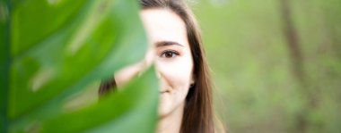 Ormanda yeşil arka plan ile büyük monstera yaprağı arkasında güzel doğal genç kadın closeup. Güzellik, sağlık ve doğa konsepti.