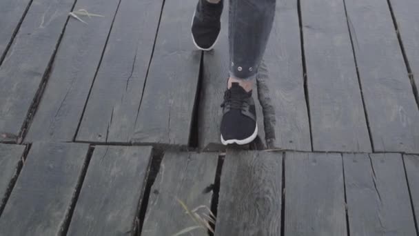Mujer en zapatillas de deporte y pantalones negros camina sobre el muelle de tablón húmedo — Vídeo de stock