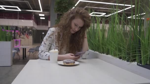 Jonge mooie roodharige vrouw in casual stijl zit in een café met koffie en donut — Stockvideo