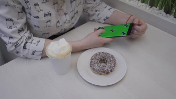 Fechar a mão de uma jovem segurando uma tela verde do telefone celular. Na mesa está um donut e café Vídeo De Stock