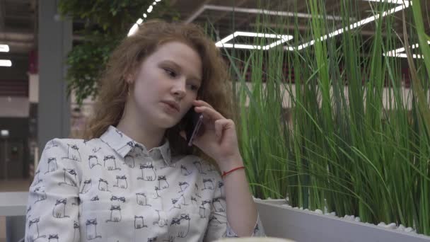 Mooie jonge vrouw zitten aan een tafel in een café, praten over de telefoon — Stockvideo