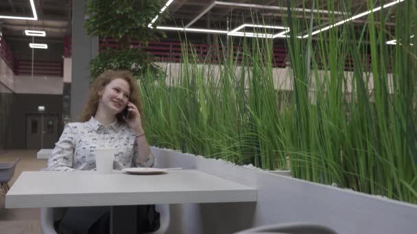 Hermosa joven sentada en una mesa en un café, hablando por teléfono — Vídeo de stock