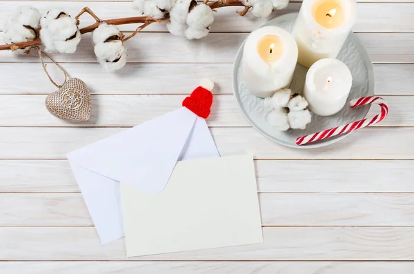 Christmas greeting card with copy space on wthite wooden background. Letter with envelope in christmas hat, candles, candy cane and cotton branch. Mock up.