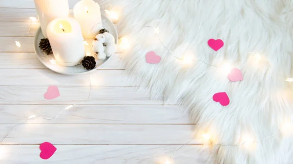 Winter cozy background for banner or greeting card. White fur, candles and hearts on white wooden background.
