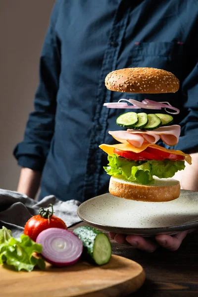 Flying burger or cheeseburger ingredients above the plate in man\'s hand on a dark background. Burger floating in the air above the table. Space for text. For banner or advertisment