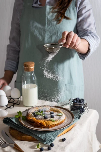 Woman\'s hands sprinkling powdered sugar on a Homemade Dutch Baby Pancake with Blueberries, mint and Powdered Sugar. Delicious Pancake, blueberries, bottle of milk and eggs on the table. Making breakfa