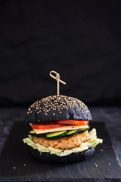 Black bun burger with delicious meat, tomatoes, cucumbers, cheese and lettuce on a black background. Side view. copy space. Restaurant, menu, fast food concept