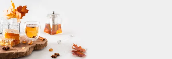 Autumn tea banner. Autumn hot tea with spices in glass cup, teapot, honey combs and falling leaves on a white background. Copy space for text, product place. Cozy fall, hygge