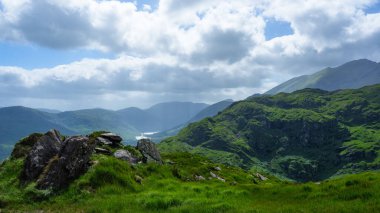 Yuvarlanan yeşil yamaçlar ve engebeli kayalar göğe yükseliyor. Uzak dağlar, İrlanda 'daki Dunloe Geçidi' nin bu sakin manzarasında sisli ufukta kaybolur..