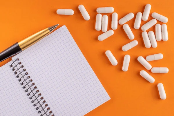 Medical capsules and pills around a blank notebook with pen on an orange background, close-up, top view, with copy space.