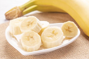Sliced and whole bananas on a white heart-shaped plate and an old wooden table in the rays of the sun. Wicker napkin. Set for healthy digestion