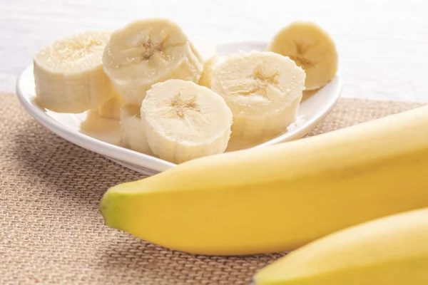 Sliced and whole bananas on a white heart-shaped plate and an old wooden table in the rays of the sun. Wicker napkin. Healthy Food Kit