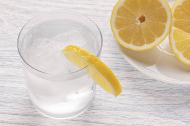 Limonlu ıslak bir bardak buz ile soğuk su yenileniyor. Beyaz ahşap bir masa üzerinde. Kullanıma hazır. Bir parça meyve beyaz tabak içinde yatıyor. Parti ferahlık içki