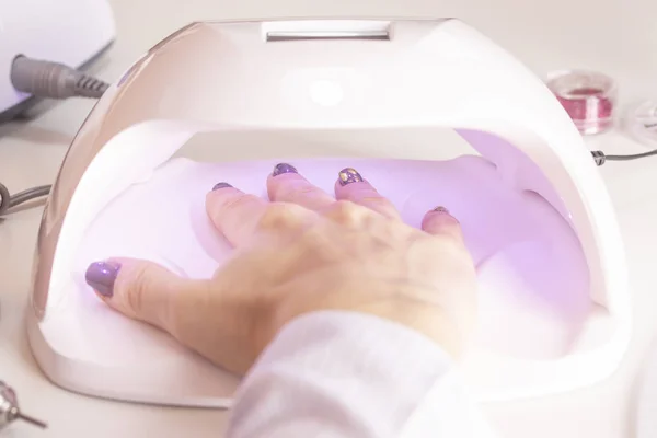 A girl's hand in an ultraviolet nail lamp with nail polish files and orange sticks of rhinestones on a manicure table in the salon