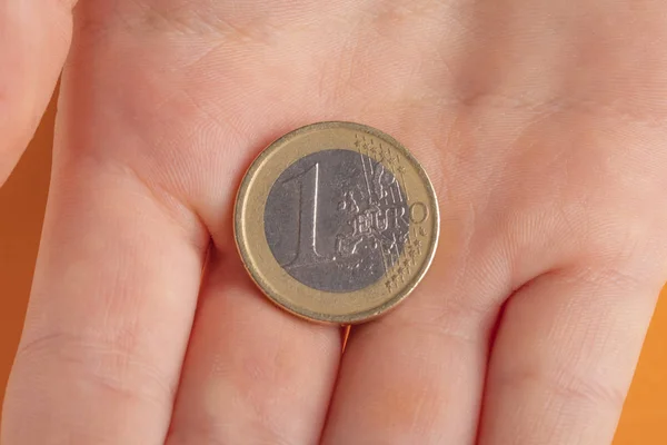 Moneda 1 euro en la mano de un niño adolescente sobre un fondo naranja . — Foto de Stock