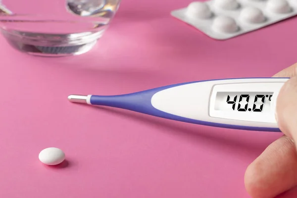 Electronic thermometer in hand and pills on a pink background. High temperature 40 degrees Celsius on display.
