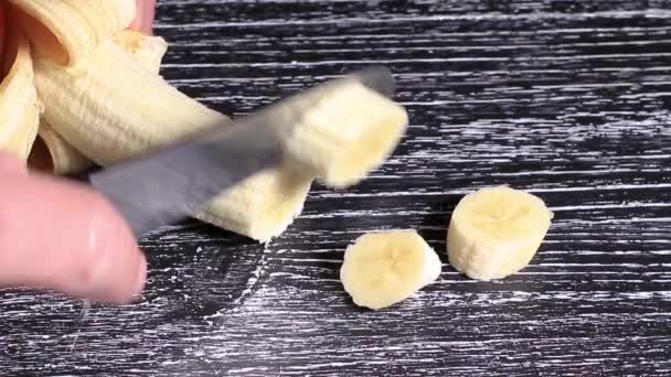 Рука человека режет спелый сочный банан на куски на черном деревянном столе . — стоковое видео
