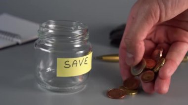 El bir etiket ve Kaydet yazıt ile bir tasarruf cam kavanoz yakınında sikke sent ve euro koyar. Not defteri, kalem, arka planda cüzdan. Gelecek için para biriktirme kavramı. yavaş çekim