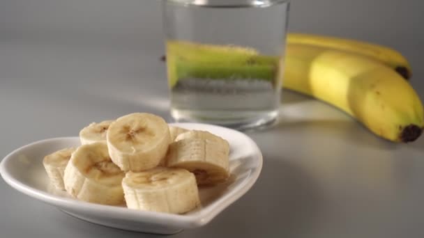 スライスし バナナ全体 白い皿の上に灰色の背景に表示されます きれいな水で霧に包まれた フルーツダイエットコンセプト — ストック動画