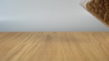 Adamın eli cam bir bardaktan tahta bir masaya bir yığın kedi maması döker ve onu yüzeye bırakır.