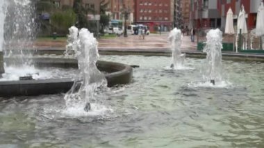 Köpüklü su akıntıları yükselir ve düşer. Şehir parkındaki çeşmeler sokağı. Yavaş çekim. Binaların, kafelerin, ağaçların ve insanların arka planına karşı