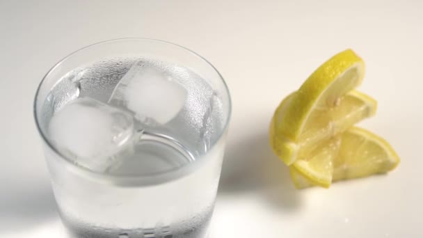 白い背景に蒸しガラスで回転する氷の立方体 次の嘘新鮮なレモンのスライス — ストック動画