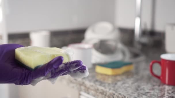 洗面台の台所で洗っていない皿の山を背景に 上から泡立つ黄緑色のスポンジに洗浄ゲルのジェットが流れます 青い保護手袋は手元にある — ストック動画