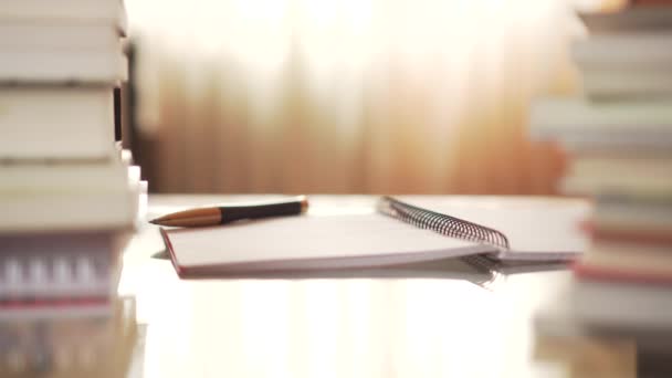 一支黑色的黄色钢笔躺在一张开着的笔记本上 放在一张玻璃桌上 在阳光照着的窗户前堆满了书 — 图库视频影像