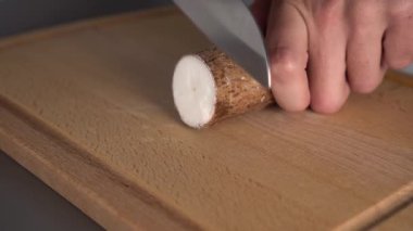 Bir adam gri bir masa üzerinde ahşap bir mutfak tahtası üzerinde bir bıçak ile yucca kökü keser. Evde egzotik yemekler pişirmek
