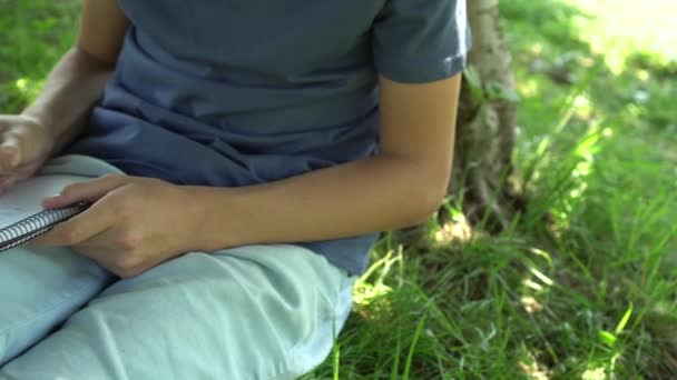 一个十几岁的少年正在树边的草地上看书 蓝色坦克上衣和鸽子短裤 黑头发考试准备 — 图库视频影像