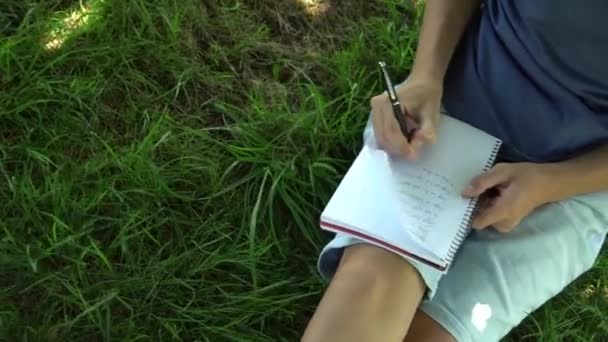 一个十几岁的少年正在树边的草地上看书 蓝色坦克上衣和鸽子短裤 黑头发 — 图库视频影像