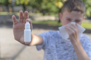 Genç çocuk burnunu darbeler ve bir kağıt beyaz mendil içine hapşırıyor ve bir parkta ağaçların bir arka plan üzerinde plastik bir kapta burun damlaları gösterir