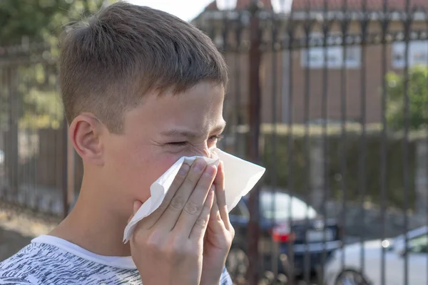 Teen boy blows his nose in a paper handkerchief on a background of fence and cars. Sneezes