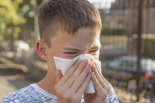 Teen boy blows his nose in a paper handkerchief on a background of trees and cars. Sneezes