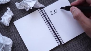 Bir erkek el gri desenli bir masa yüzeyine 2020 gol atar. Sayfayı yırtıp atıyor. Buruşmuş kağıtlar yakınlarda. Yeni yılda planlama için karmaşık yaratıcı arayış kavramı