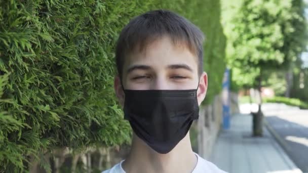 少年笑了笑 并戴上了一个医疗保护的黑色面具 在城市街道上 道路旁边有绿色的植被 大肠癌流行与过敏防护措施 — 图库视频影像