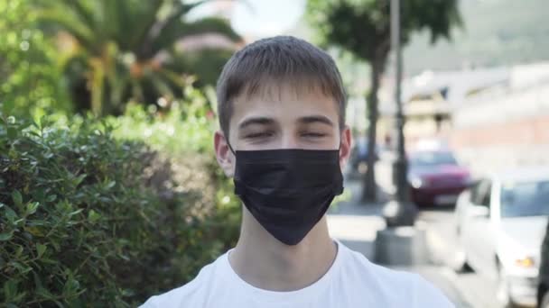 年轻人笑了 并戴上了一个医疗保护黑色面具在城市街道上与棕榈树和绿色植被旁边的道路 大肠癌流行与过敏防护措施 — 图库视频影像