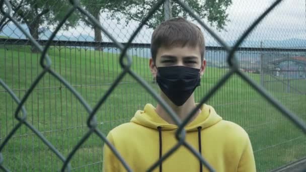 若い男は鉄のワイヤーフェンスの後ろに医療用の黒いマスクを脱いだ コロナウイルスのパンデミックと制限措置からのうつ病の概念 — ストック動画