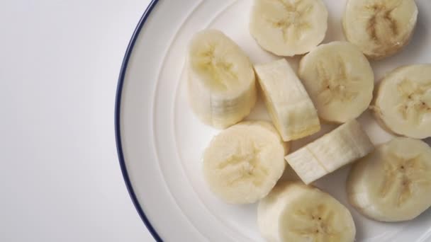 青い境界線のクローズアップと2つの皮をむかないバナナの白いプレート上の熟したバナナのスライス 回転が遅い — ストック動画