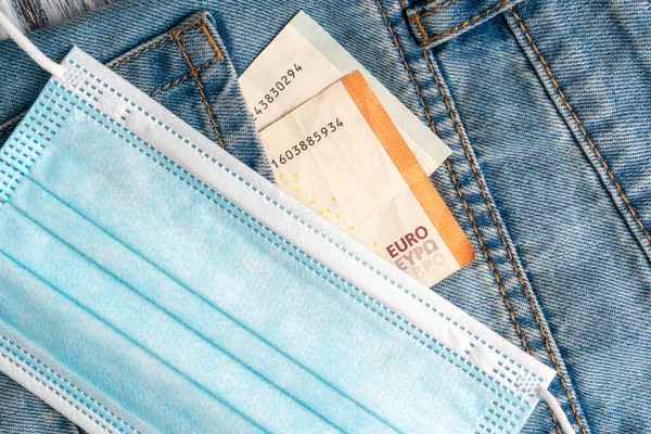 Gri ahşap bir arka planda mavi eski kot pantolon cebinde koruyucu bir tıbbi maske ve euro banknotları yakın plan. Küresel mali kriz ve işsizlik kavramı