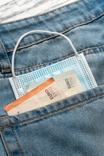 Close-up of a protective medical mask and euro banknotes in the pocket of blue old jeans on a gray wooden background. The concept of the global financial crisis and unemployment. Vertical view