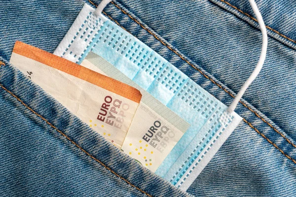 Close-up of a protective medical mask and money in the pocket of old jeans on a wooden background. Global financial crisis and unemployment concept