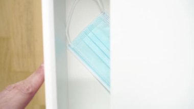 El, komodinin rafını tıbbi koruyucu bir maskeyle kapatıyor. Coronavirus 'a karşı günlük kişisel koruyucu ekipman. Kahverengi parke üzerinde beyaz mobilyalar. Seçici odak
