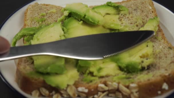 健康的なアボカドサンドイッチを作る ナイフのクローズアップは 熟した柔らかいアボカドの層をシリアルパンに広げます 暗い石の背景に白いプレート — ストック動画