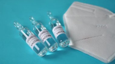Mavi bir yüzeyde COVID-19 koronavirüs aşısı ampul ve solunum maskesi. Koronavirüs enfeksiyonunun önlenmesi, aşılanması ve tedavisi için