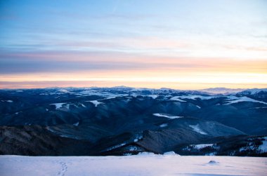 Karlı dağlar bulutlu günbatımı gökyüzünün altında görünür.