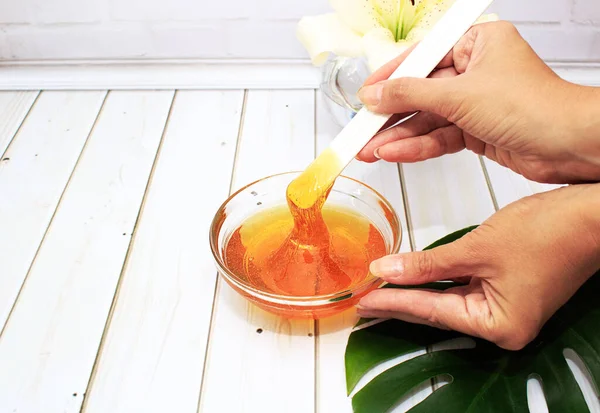 Femal hands hold liquid sugar paste for hair removal in a bowl with lily flower and dracaena leaf in glass vase. The concept of body care, beauty treatments.